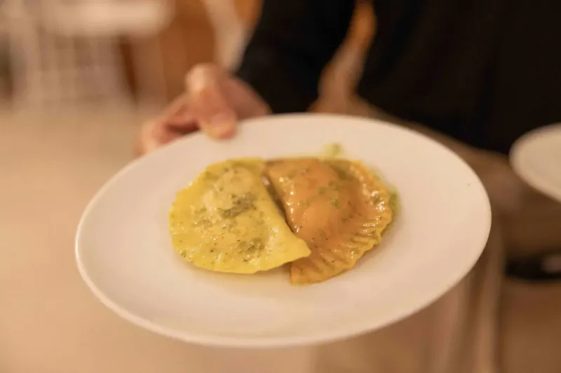 Hand holding white plate with two ravioli: one yellow with green herbs, one orange sauce, blurred restaurant background.