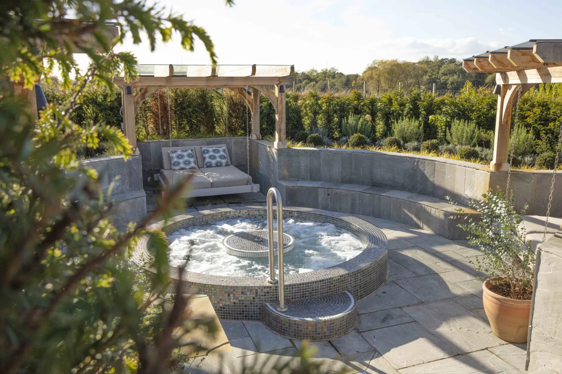 Bubbling hot tub in landscaped garden with pergola, cushioned benches, surrounded by greenery at spa resort.