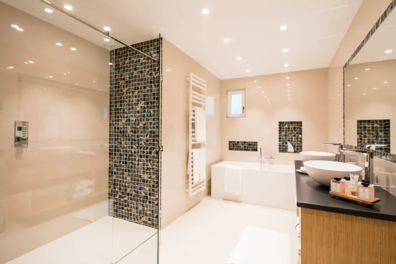 Modern luxury hotel bathroom with glass shower, mosaic tiles, freestanding tub, and warm lighting.