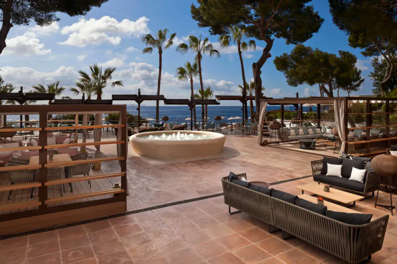 Outdoor terrace at ARRELS by Marga Coll with central fire pit, lounge seating, palms, pines, and sea view.