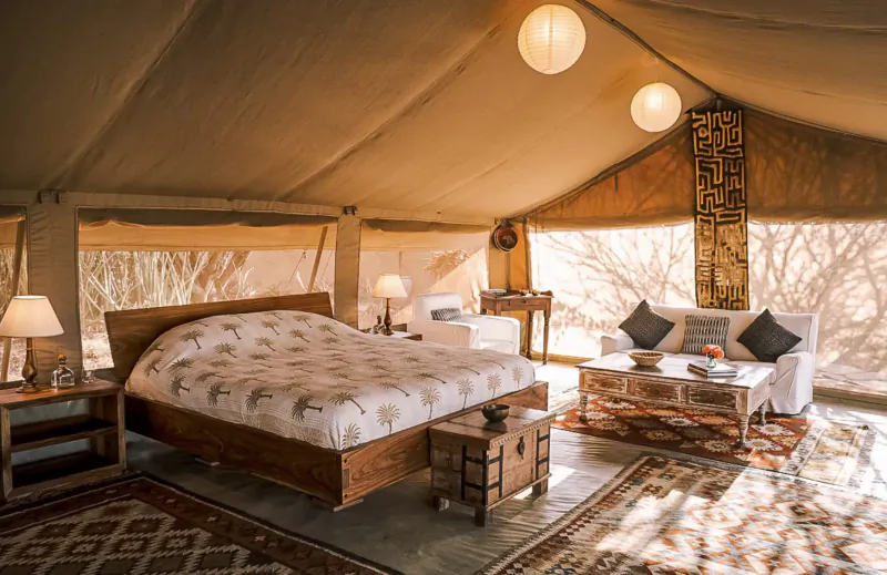 Luxurious tent interior at Tumaren Camp with wooden bed, white pillows, low table, sofa, lamps, and desert view.