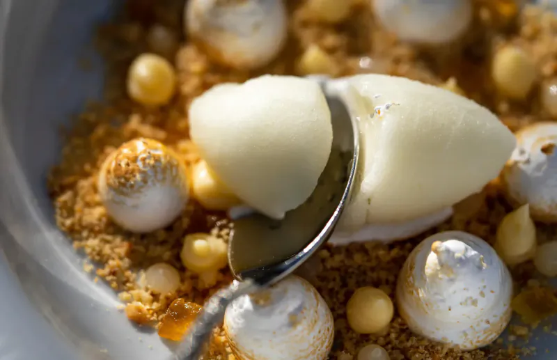 Close-up of dessert in bowl: white ice cream scoops, meringues, white beans, golden crumble, spoon scooping ice cream