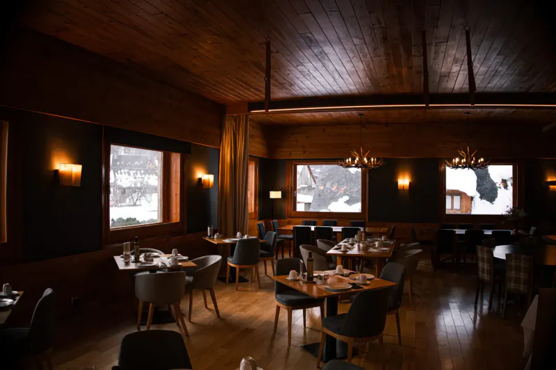 Cozy wooden ski chalet dining room with tables, chairs, snowy mountain views through windows.