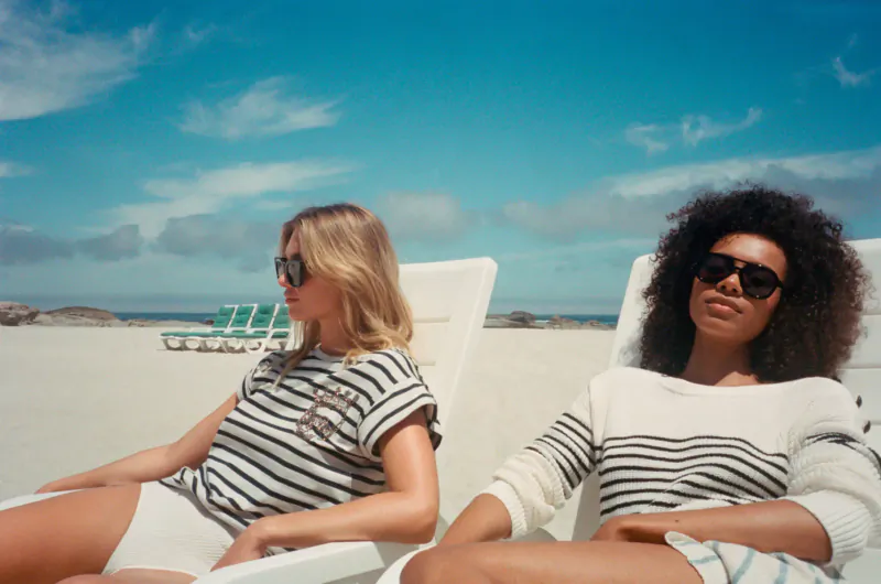 Two stylish women in striped sweaters and sunglasses lounging on white chairs on sunny beach.