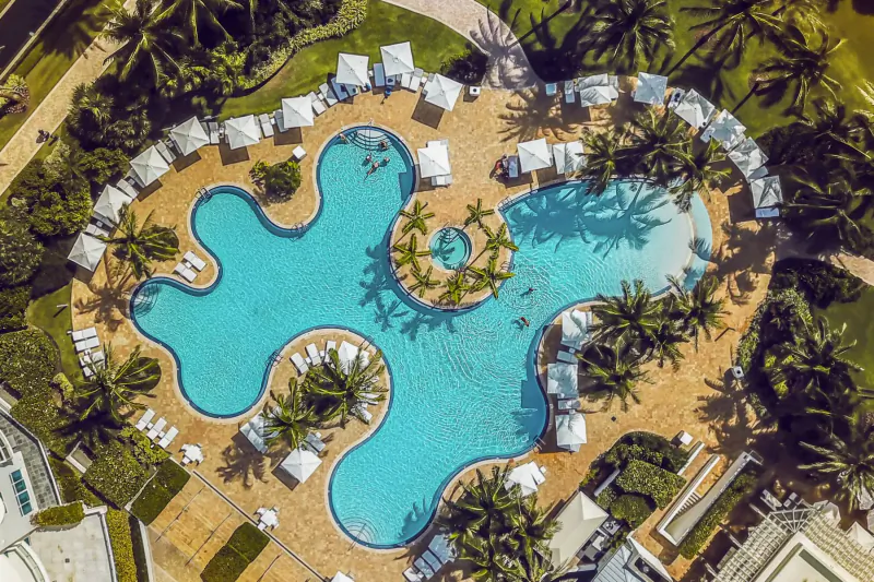 Aerial view of Continuum's curvy turquoise pool surrounded by white loungers, palm trees, and green lawns at the sporting club spa.
