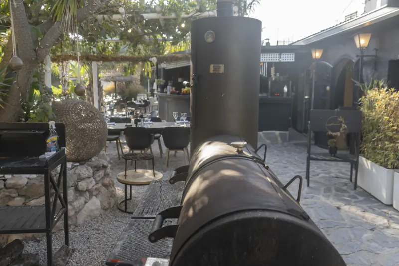 Close-up of large black smoker barbecue in La Vuelta Boutique Hotel's tropical outdoor courtyard with tables and plants.