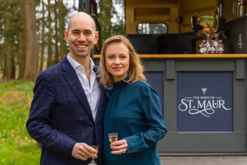 Smiling bald man in navy suit and woman in teal dress hold St. Maur liqueur glasses by branded bar in woods.