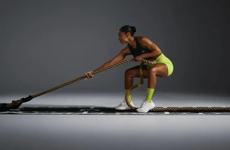 Fit woman in black sports bra and lime green shorts pulling thick rope in gym workout, squatting dynamically.