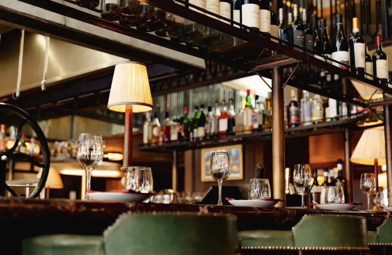 Elegant Italian steakhouse bar with wooden shelves of wine bottles, green stools, wine glasses, and warm lamps.
