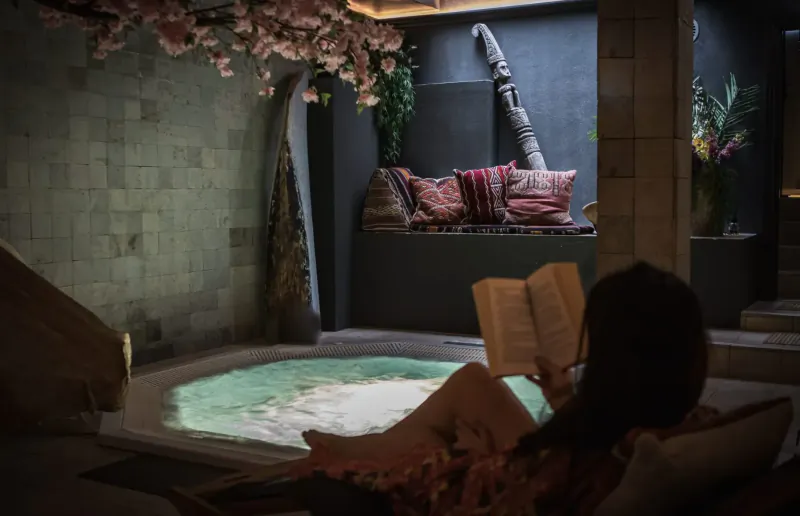 Woman relaxing in green-lit hot tub at Manon Les Suites, reading book amid hanging cherry blossoms and lush plants