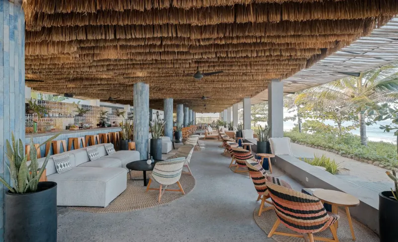 Open-air dining lounge at Amber Lombok with thatched roof, woven chairs, plants, and ocean views.