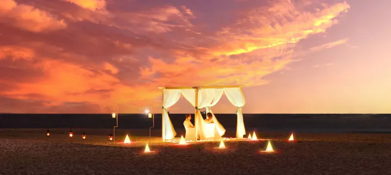 Bride and groom under white-draped beach gazebo at sunset, surrounded by candles on sand during Eid celebration