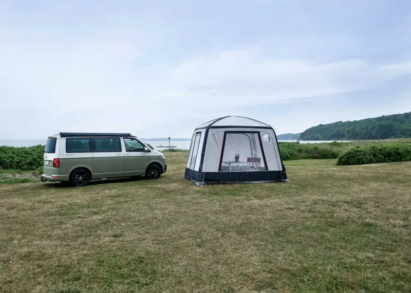 Green VW camper van parked on grassy field beside white Isabella awning tent, beach in background.