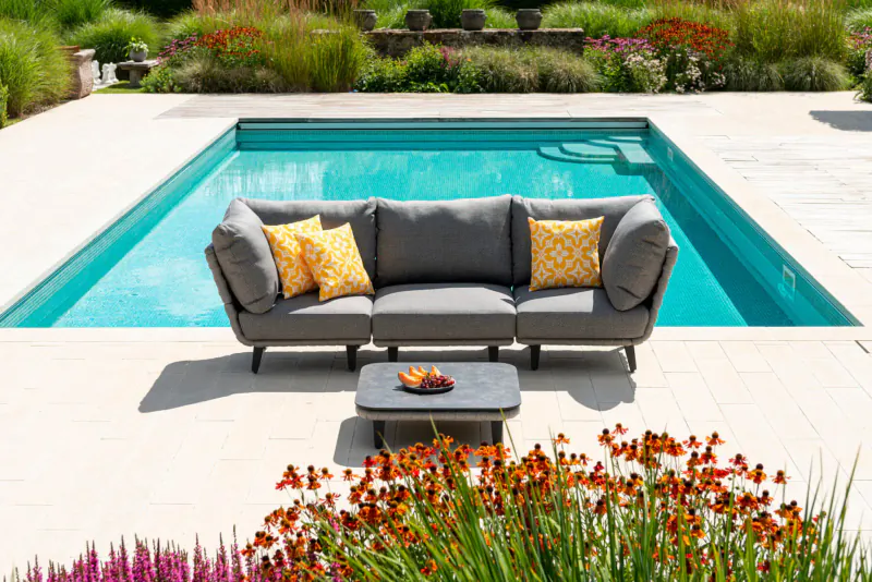 Gray Alexander Rose outdoor sofa with yellow cushions and low table with fruit, beside turquoise pool in lush garden.