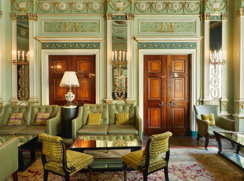 Elegant Home House club interior with ornate green walls, wooden doors, green velvet sofas, armchairs, lamps, and mirrors.