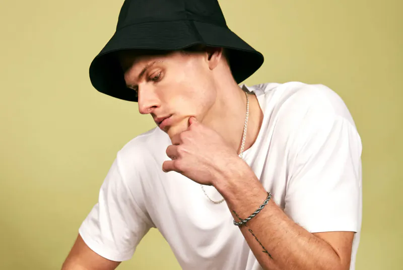 Man in black bucket hat and white tee, hand on chin thoughtfully, yellow background, sustainable menswear.