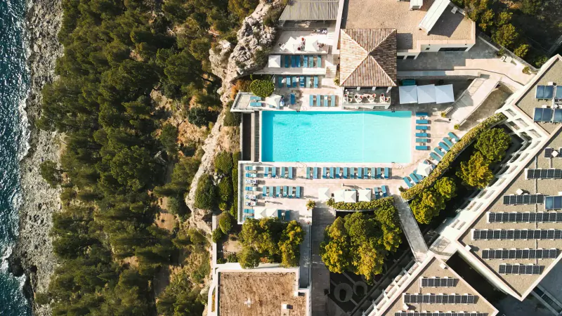 Aerial view of Jumeirah Mallorca's infinity pool overlooking the sea, with lounge chairs, umbrellas, and lush gardens.