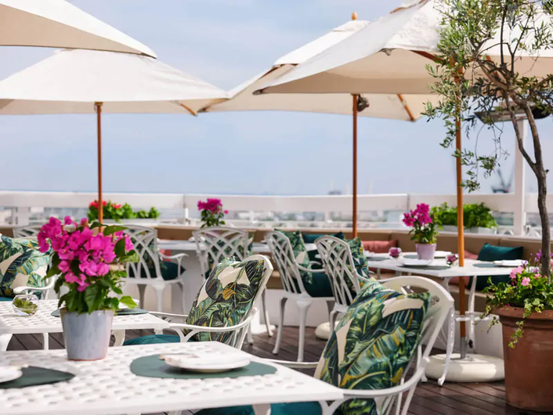Elegant rooftop al fresco dining at The Dorchester: white umbrellas, green patterned chairs, pink flowers, city skyline.