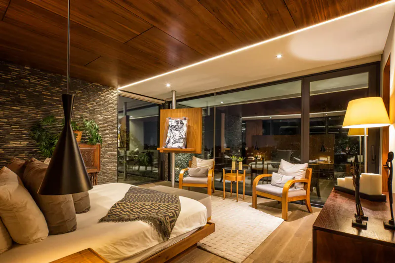 Modern bedroom with wooden ceiling, stone walls, large window, bed, chairs, plants, and abstract art.