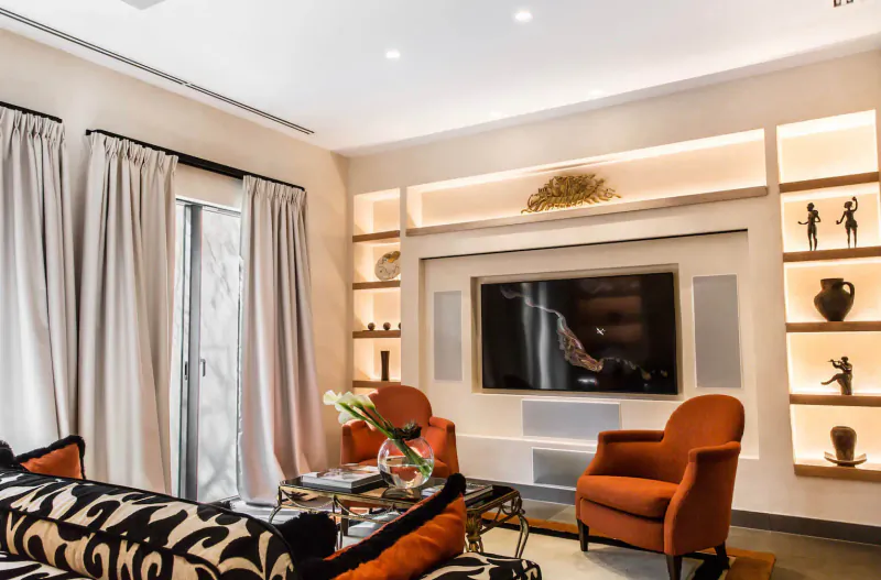 Elegant living room in Terre Blanche Provence oasis with orange armchairs, zebra sofa, lit shelves with vases and sculptures, large TV.