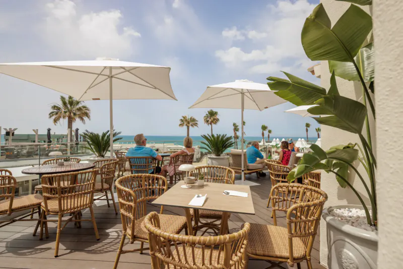 Outdoor terrace restaurant at Vale do Lobo resort with wicker tables, umbrellas, plants, guests, and ocean-palm view.