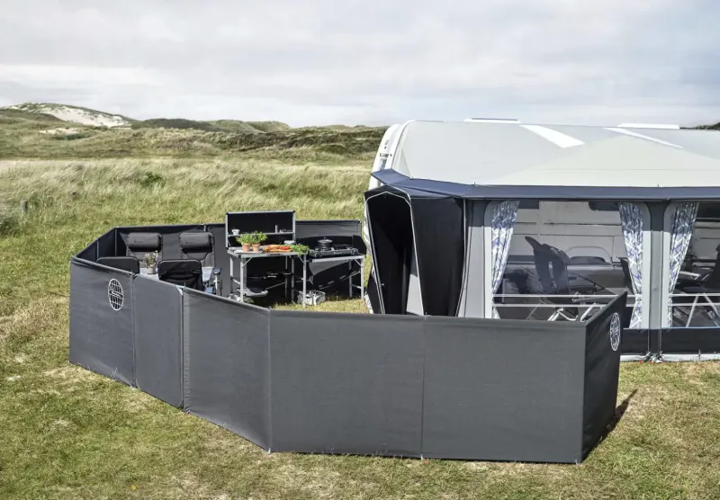 Black modular privacy fence with kitchen setup and chairs beside luxury campervan on grassy dunes under cloudy sky.