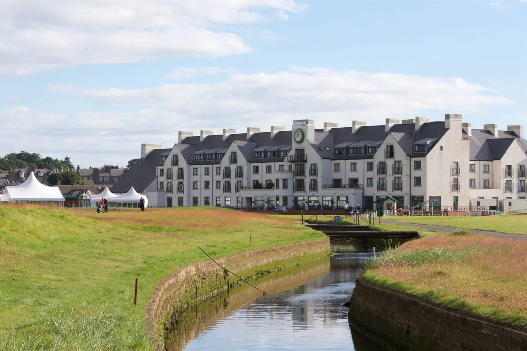 White Carnoustie golf resort with green clock tower, tents, and stream on golf course under blue sky