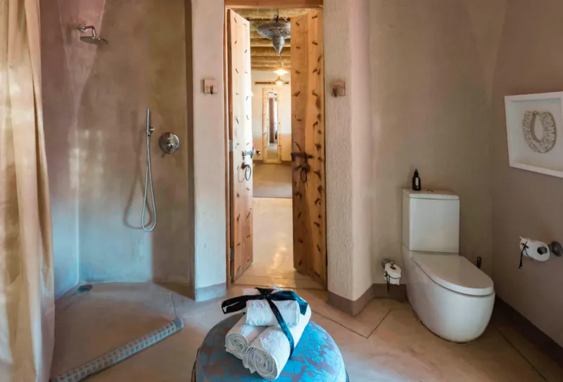 Spacious rustic ensuite bathroom with open shower, white toilet, blue stool with towels, and door to bedroom.