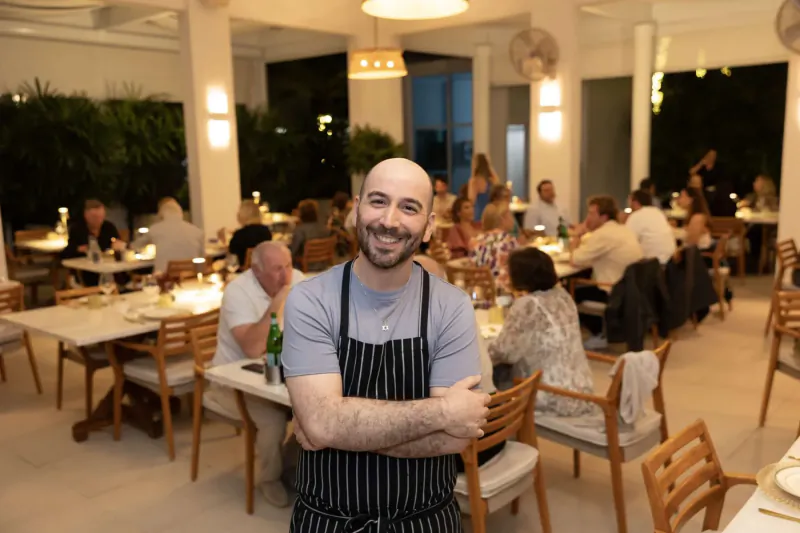 Smiling bald chef in black apron arms crossed, standing in busy upscale restaurant dining room