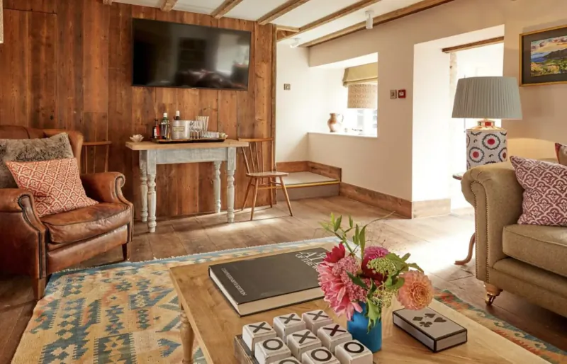 Cozy Cotswolds inn lounge with wooden beams, leather armchairs, floral vase, coffee table with games and book, TV on wood wall.