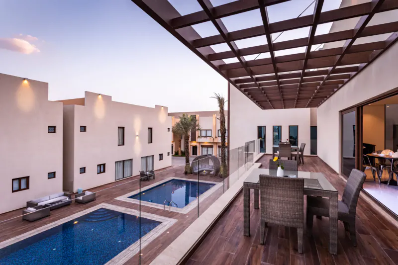 Luxury villa resort terrace at Braira Hattien with infinity pools, wooden dining table, lounge chairs, and palm trees at dusk.