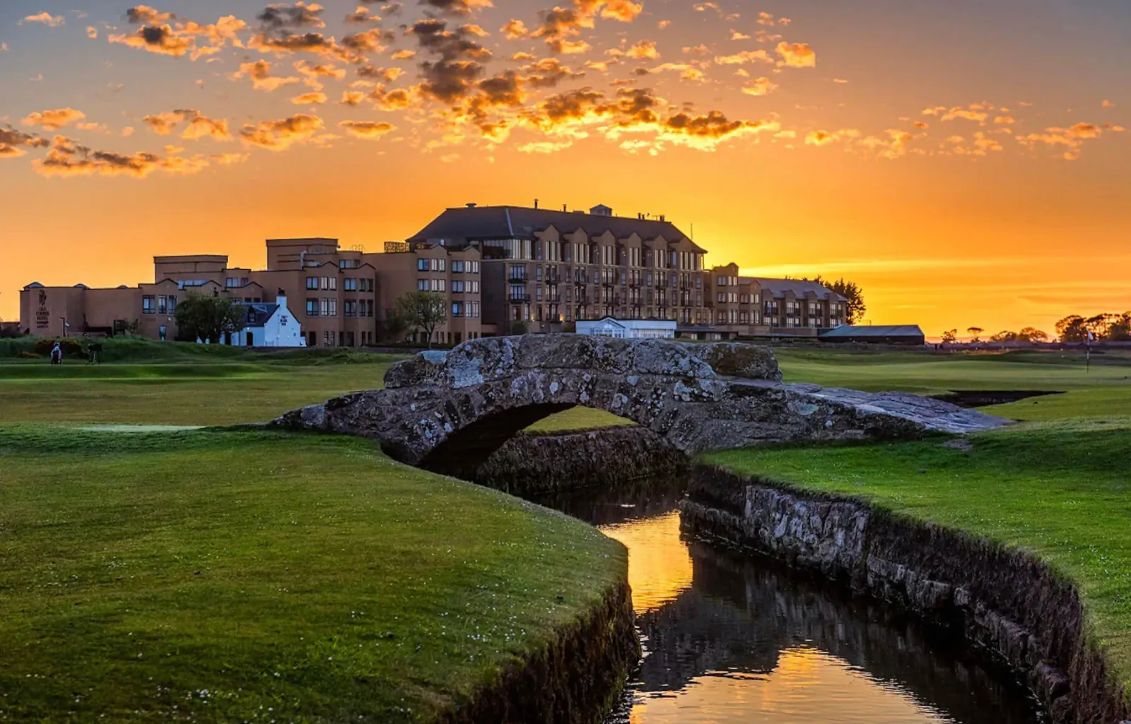 Stone bridge over stream on golf course at sunset, with historic hotel and orange sky behind.