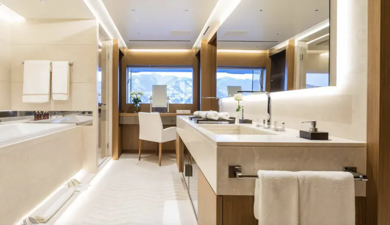 Luxurious yacht bathroom with white marble vanity, chair, towels, flowers, and alpine window view.