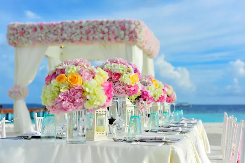 Elegant beach wedding table with colorful floral arrangements under a flower-draped canopy, ocean backdrop.