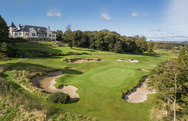 Aerial view of The Old Course Hotel overlooking manicured golf course with bunkers and fairways under blue skies.
