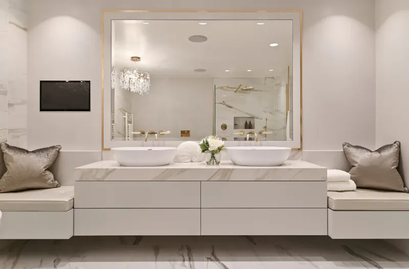 Elegant modern bathroom with large gold-framed mirror, white marble double vanity, chandelier, and white roses