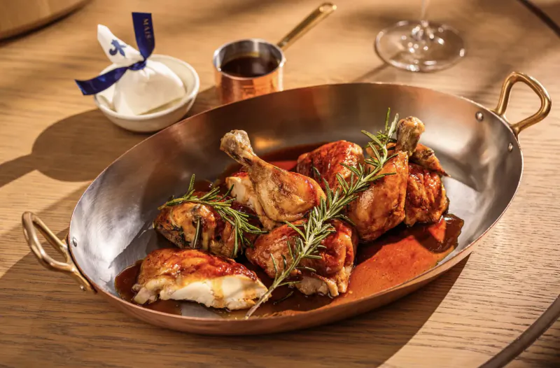 Roasted chicken legs with rosemary in copper pan, garnished with blue napkin, on wooden table at upscale dining setting.