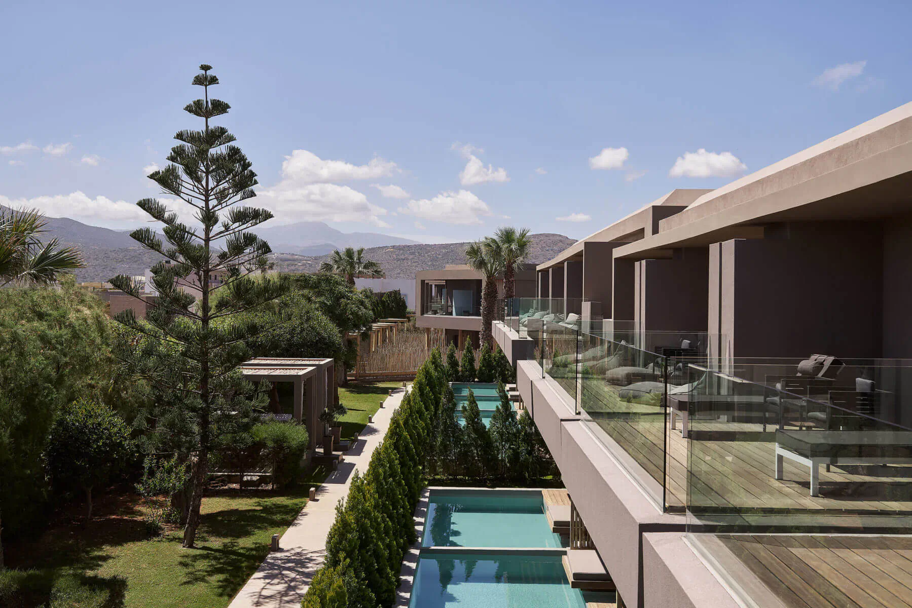 Aerial view of Elysium Boutique Hotel Crete: modern terraced suites with glass balconies, turquoise pools, palm trees, gardens, and distant mountains under blue sky.