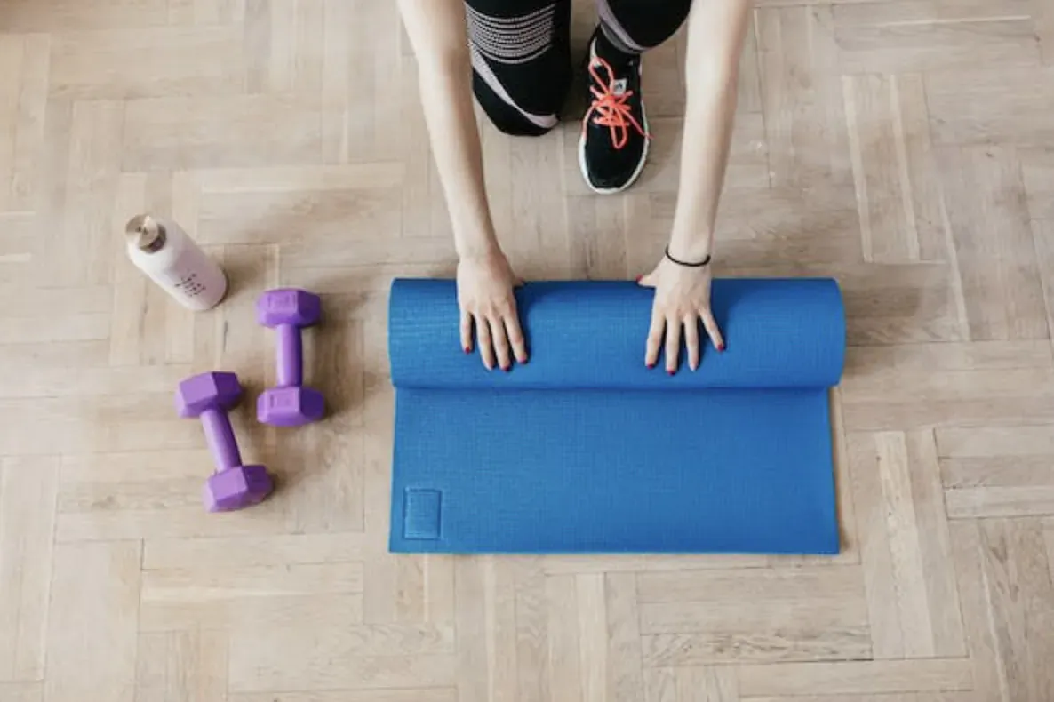 Overhead view of woman kneeling in red sneakers, unrolling blue yoga mat beside purple dumbbells and water bottle on wood floor