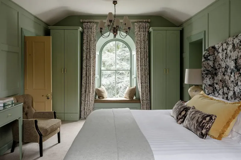 Elegant green bedroom in Boyne Valley with arched window seat, floral headboard, chandelier, and armchair.
