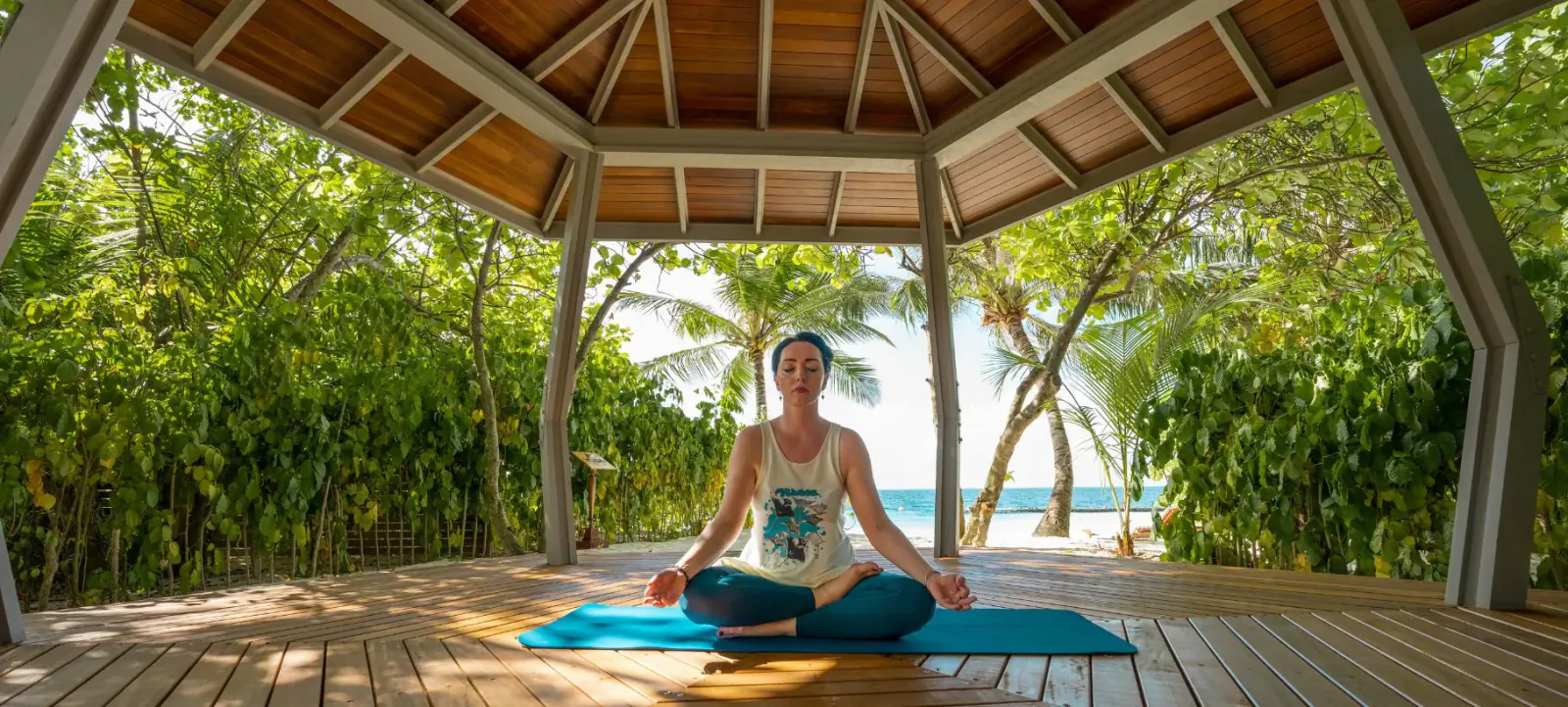 Woman in headscarf meditating in lotus pose on blue yoga mat in open wooden beach gazebo surrounded by tropical greenery and ocean.
