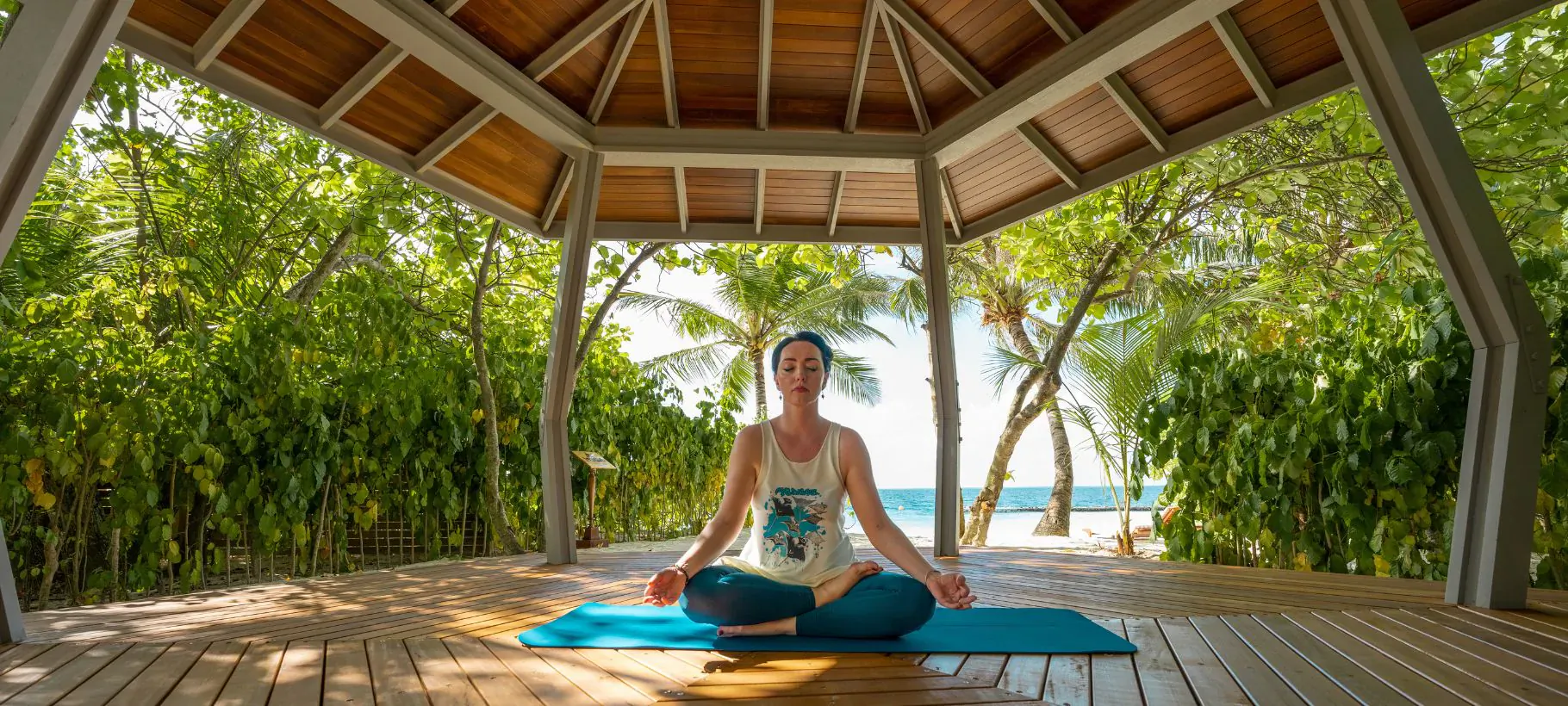 Woman in headscarf meditating in lotus pose on blue yoga mat in open wooden beach gazebo surrounded by tropical greenery and ocean.