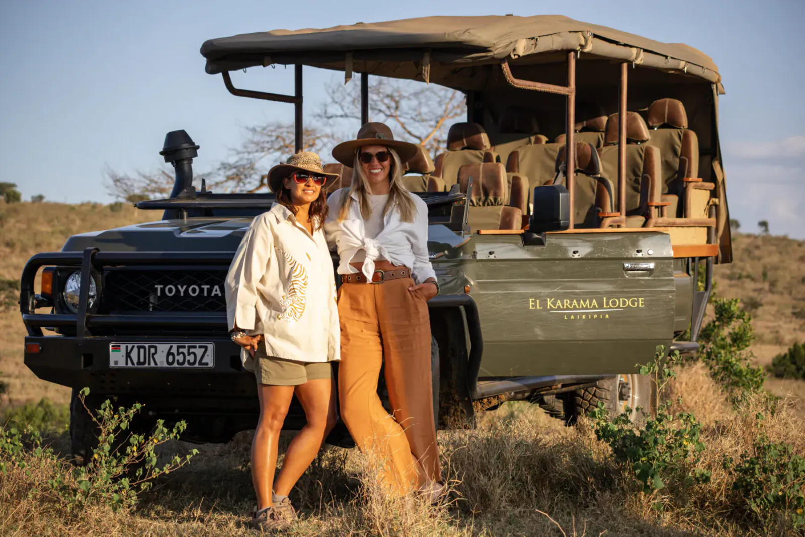 Two women in safari outfits stand smiling by green El Karama Lodge Land Cruiser in African bush, license KOR 655Z.