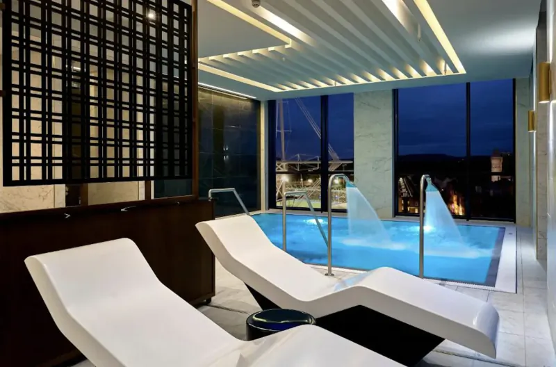 Modern indoor infinity pool with glowing blue water, white lounge chairs, and city night view through windows at The Parkgate Hotel.