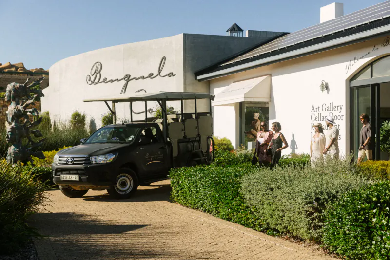 Benguela Cove building with 'Benguela' sign, Art Gallery, parked black Toyota Hilux truck, people outside amid greenery
