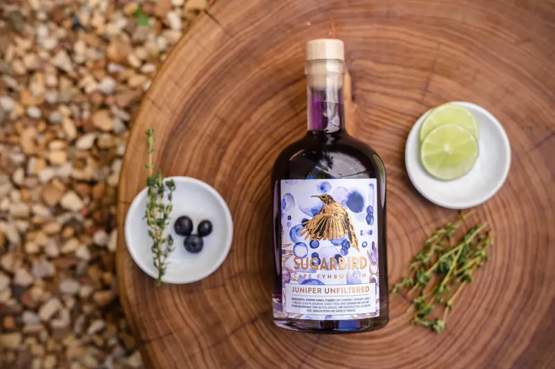 Sugarbird Cape Fynbos Gin bottle with bird label on wood table, surrounded by blueberries, limes, and thyme on pebbles.