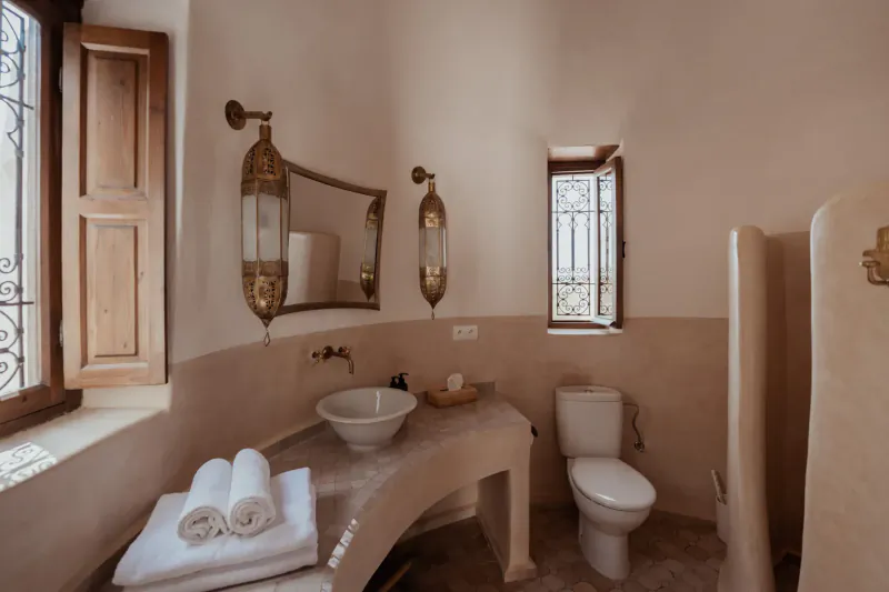 Elegant Moroccan-style bathroom in Les Jardins De Villa Maroc, with arched windows, brass lanterns, curved sink, towels, and shower.
