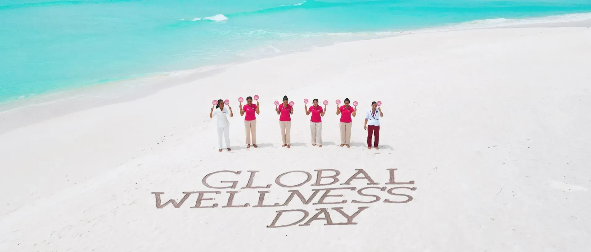 Six women in red and white outfits stand smiling on white sand beach by turquoise sea, with 'GLOBAL WELLNESS DAY' written in sand.