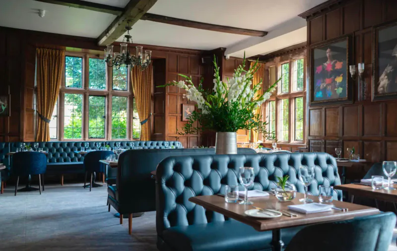 Elegant lounge at Billesley Manor Hotel with dark wood paneling, blue velvet sofas, large floral arrangement, and tall windows.