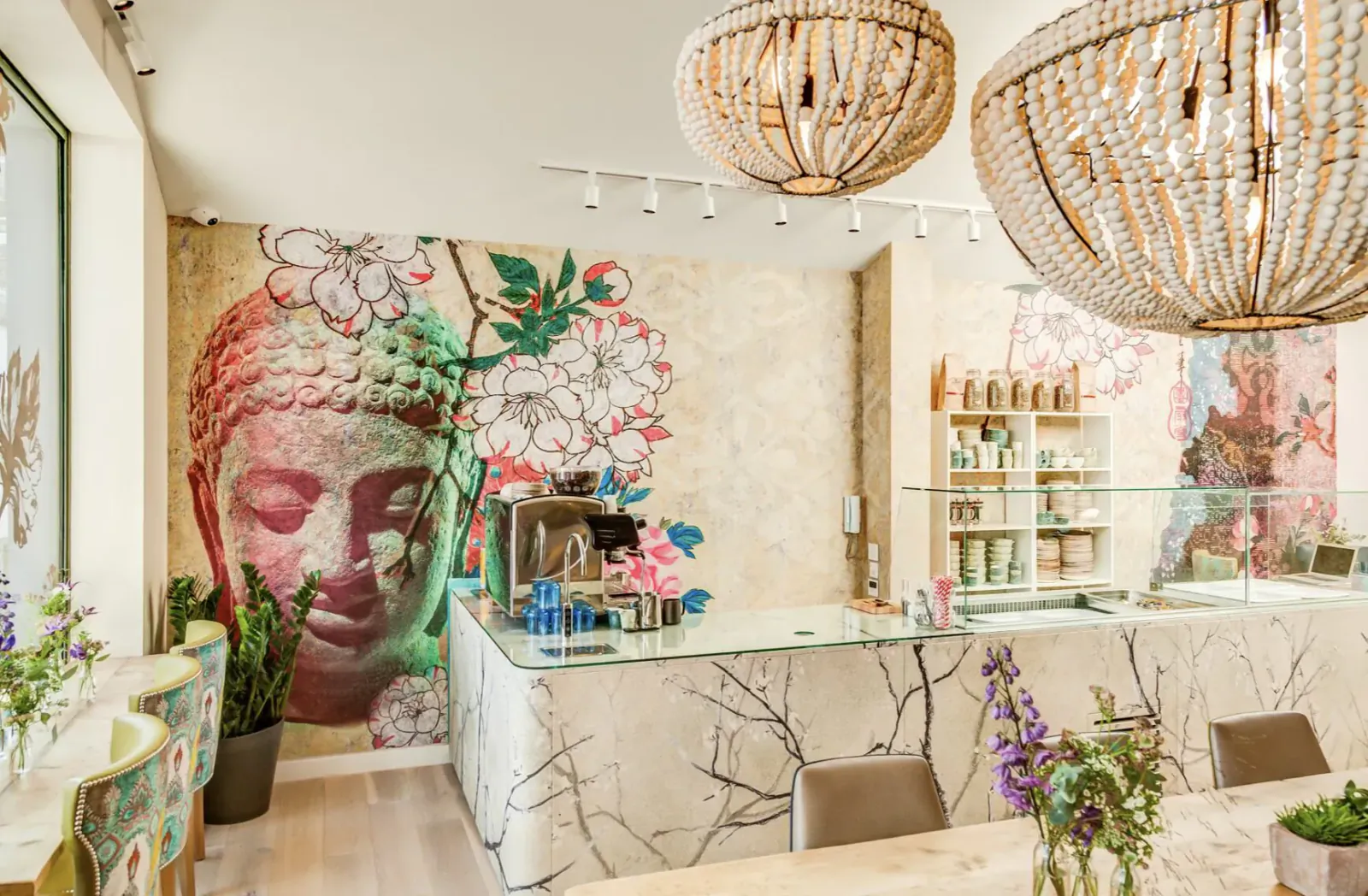 Cozy wellness cafe interior with large pink Buddha mural, rattan pendant lights, potted plants, and wooden counter.